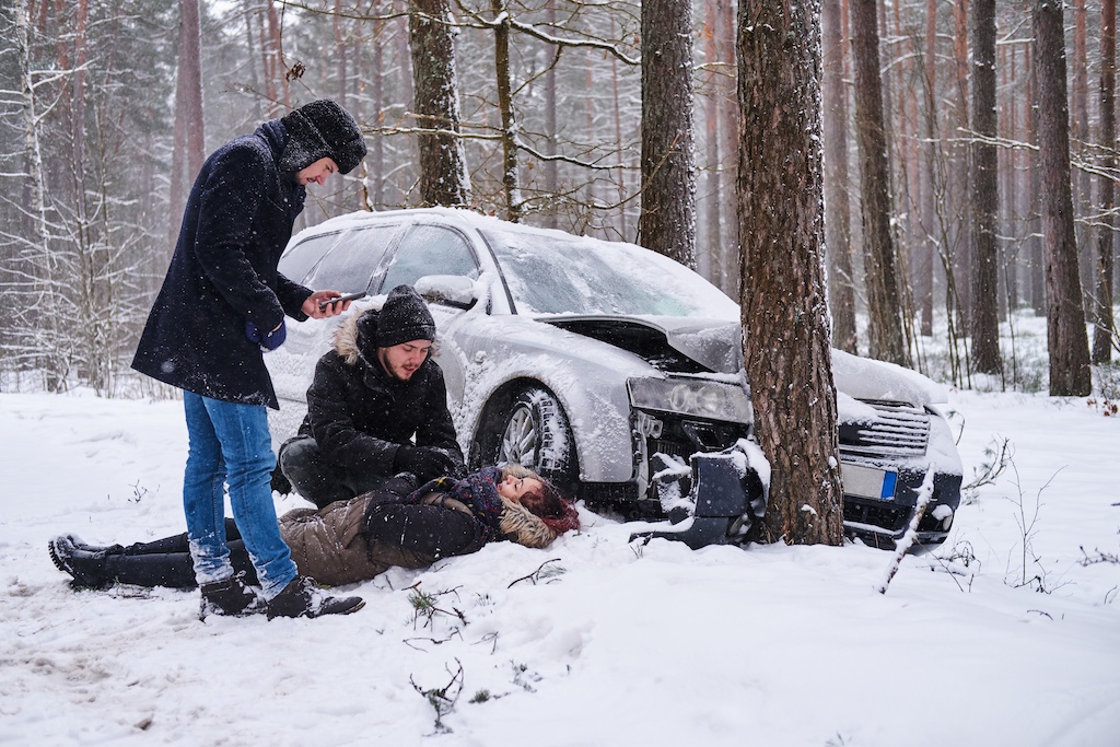 Two people in winter coats are providing emergency assistance to an injured woman lying in the snow beside a damaged white car after a car accident injury.