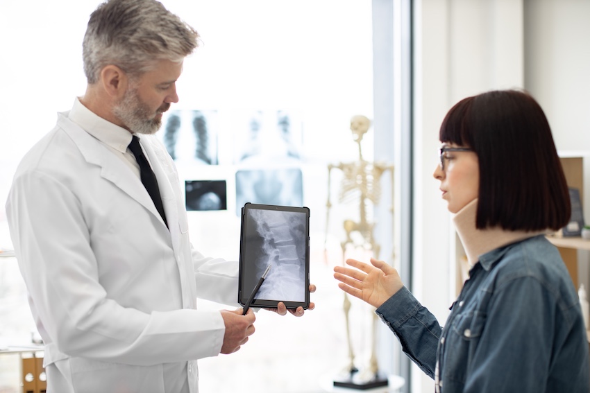 A male doctor with short silver-gray hair and a neatly trimmed beard wearing a white lab coat over a black dress shirt and tie holds a black tablet displaying a detailed spinal X-ray image and points to it with a stylus while a female patient with a dark bob haircut- glasses- and a white cervical neck brace gestures with an open hand as she describes her symptoms both standing in a bright clinical examination room with multiple additional X-ray images mounted on a lightbox in the background and a full anatomical skeleton model visible on a shelf to the right — illustrating the spinal imaging review and diagnostic consultation a car accident doctor conducts with an injured patient in Georgia.