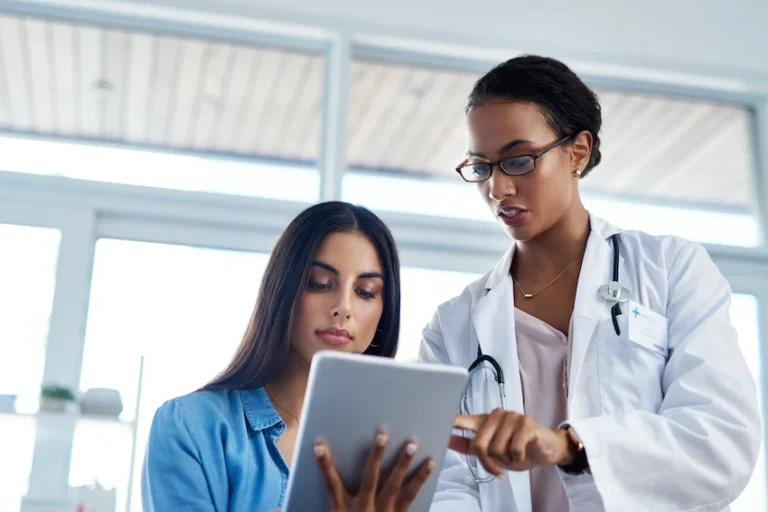 A female doctor in a white lab coat and stethoscope points to information on a tablet while a young female patient looks on attentively illustrating the type of detailed medical review and injury assessment a car accident doctor conducts with patients following a collision in Georgia.