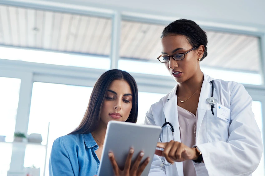 A female doctor in a white lab coat and stethoscope points to information on a tablet while a young female patient looks on attentively illustrating the type of detailed medical review and injury assessment a car accident doctor conducts with patients following a collision in Georgia.