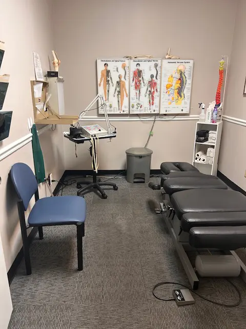 A chiropractor's exam room with a table, chair, posters of a spine, and equipment