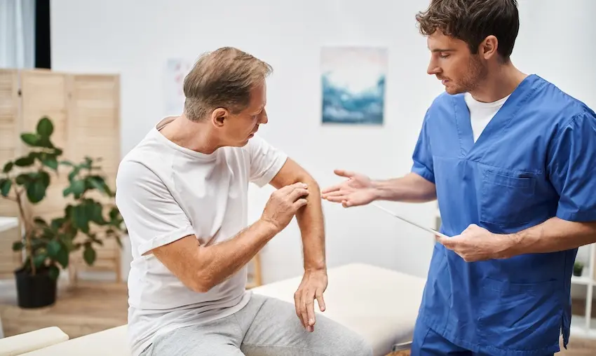 A middle-aged man in a white t-shirt sits on a medical exam table pointing to his shoulder while a young male doctor in blue scrubs from 1800hurt911ga.com holds a tablet and gestures attentively illustrating the clinical evaluation process used to diagnose hidden injuries that may not have been apparent at the scene of an accident.