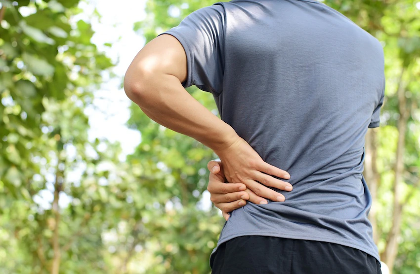 A man in a gray t-shirt and black shorts stands outdoors among green trees pressing both hands firmly against his lower back in pain representing the delayed onset of hidden injuries like lumbar strain and soft tissue damage that accident victims commonly experience days after a collision in Georgia and could use help from 1800hurt911ga.com.