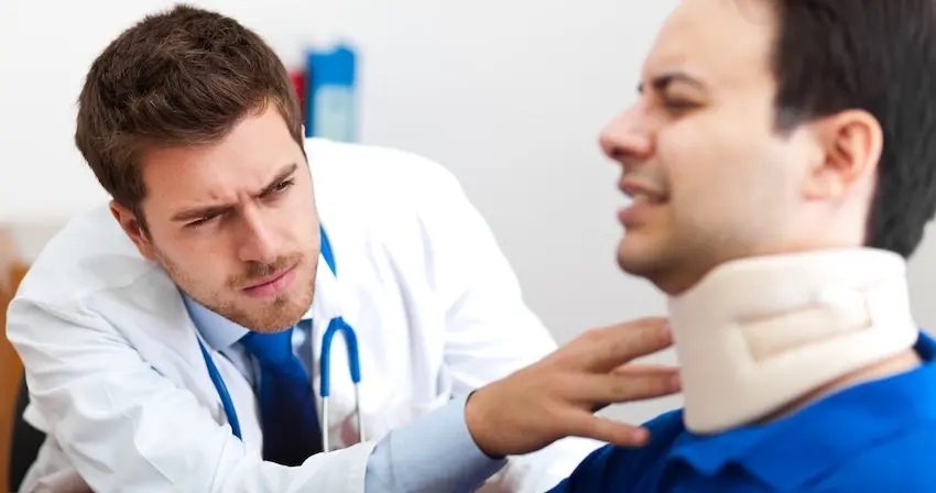 A young doctor in a white lab coat carefully examines the neck of a male patient wearing a cervical collar depicting the hands-on physical evaluation a physician performs to assess the severity of car accident and whiplash injuries sustained in a collision in Georgia.