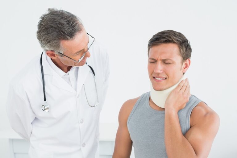 An older doctor with glasses leans in to observe a young male patient in a neck brace who winces and holds the side of his neck in pain illustrating the clinical examination process used to diagnose and document car accident and whiplash injuries with help from 1800hurt911ga.com before beginning treatment.