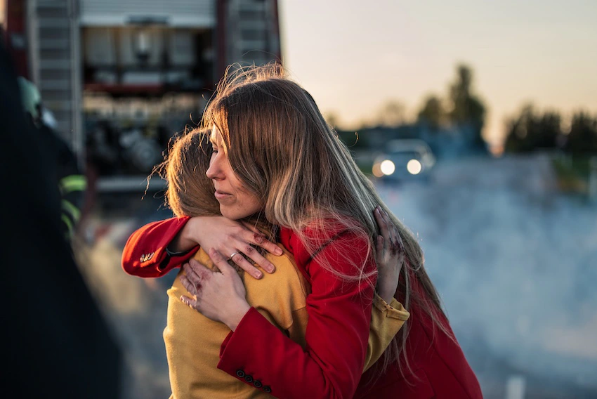 A woman in a red jacket with long blonde hair embraces another person in a yellow jacket in an emotional hug at dusk with smoke- emergency vehicles- and blurred lights visible in the background- capturing the raw grief and shock that car accident victims and their loved ones experience in the immediate aftermath of a serious crash in Georgia.