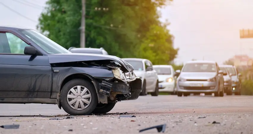 A black sedan with a heavily crumpled front bumper and scattered debris on the pavement sits stopped on a city road at golden hour with a line of cars with headlights on backed up in the background depicting the chaotic traffic scene that car accident victims are left to manage after a collision in Georgia.
