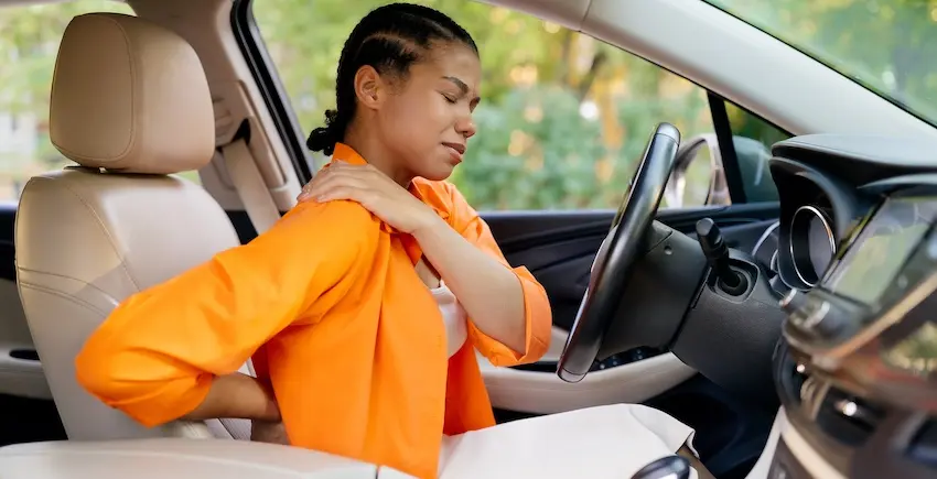 A woman in a bright orange shirt sits in the driver's seat of a car with her eyes closed reaching one hand back to grip her neck and the other pressing against her lower back illustrating the neck and back pain that car accident victims commonly experience immediately following a rear-end collision and could use help from 1800hurt911ga.com.