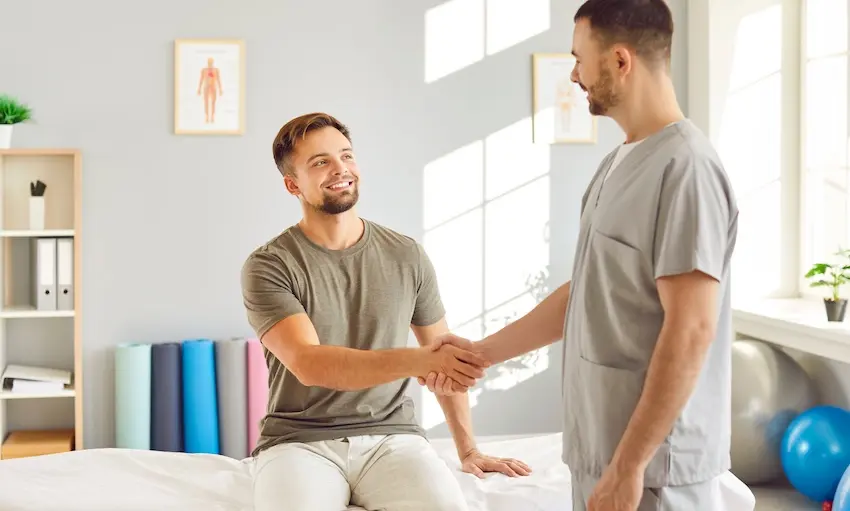 A smiling young bearded man in an olive green t-shirt sits on a padded treatment table and shakes hands with a male chiropractor from 1800hurt911ga.com in gray scrubs in a bright rehabilitation room with exercise mats, anatomy charts, and therapy equipment visible in the background, representing the encouraging progress car accident victims can make when connected with proper medical care and treatment in Georgia.